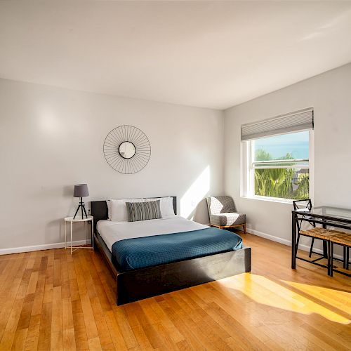 A modern, bright room with a bed, bedside table, wall decor, chair, window, and small dining table on a wooden floor.