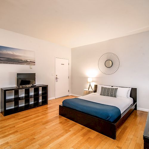 A modern bedroom with a bed, TV on a stand, a fan, a chair, a lamp, and wall art on light-colored walls.