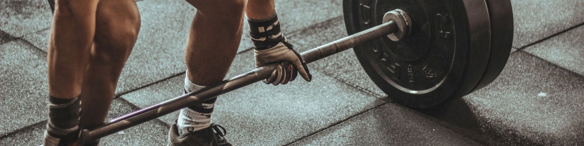 A person in sports attire is preparing to lift a heavy barbell in a gym setting. The gym floor is covered in rubber mats.