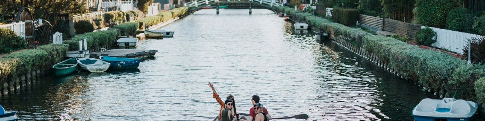 A couple is rowing a boat in a tranquil, narrow canal, surrounded by lush greenery and small boats along the banks.