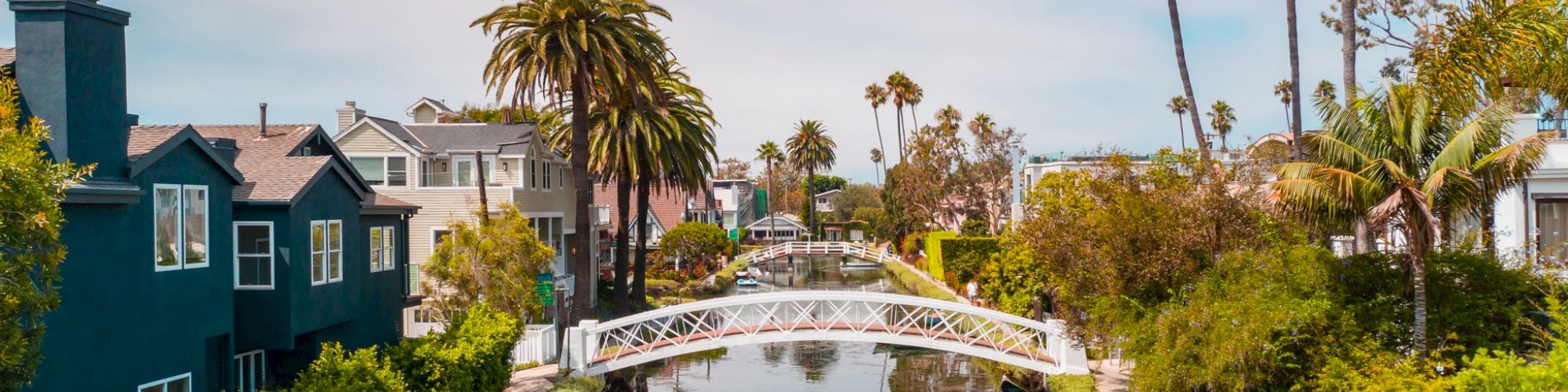 A scenic canal lined with houses, palm trees, and lush greenery, featuring a small white footbridge and a red canoe floating in the water.