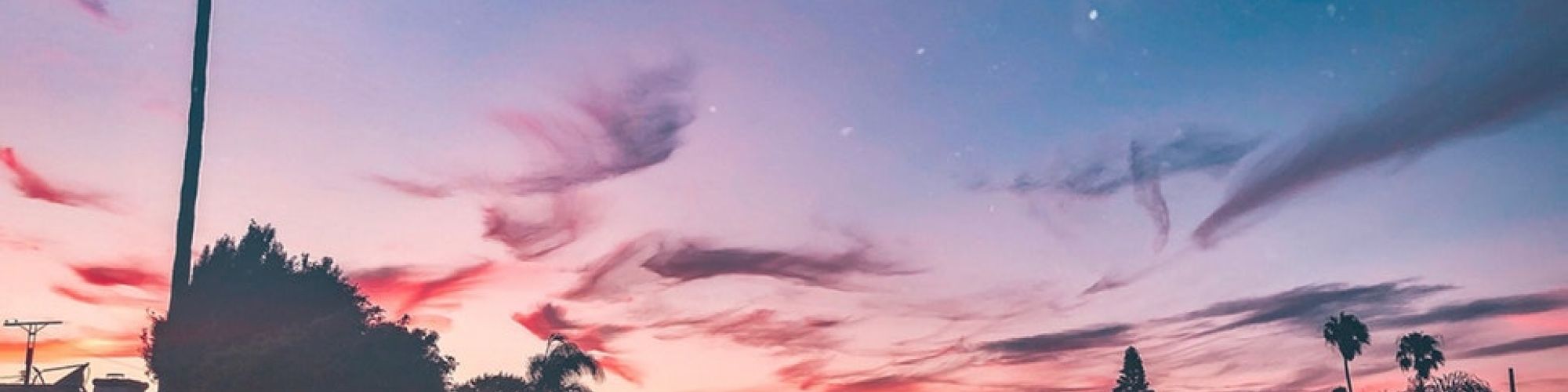 A tranquil scene of a sunset or dawn over a canal with silhouetted palm trees, houses, and a colorful sky filled with clouds and stars.