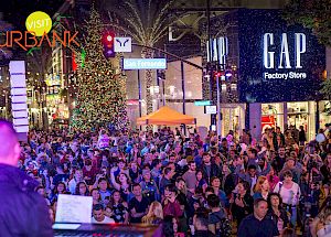 A large crowd is gathered on a street near a GAP Factory Store. It appears to be a festive event with lights and decorations, including a Christmas tree.