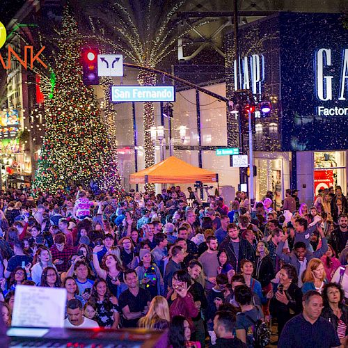 A large crowd is gathered on a street near a GAP Factory Store. It appears to be a festive event with lights and decorations, including a Christmas tree.