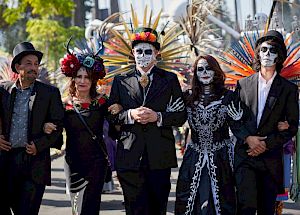 A group of people dressed in festive costumes with face paint, possibly celebrating a cultural festival, walking together arm in arm.