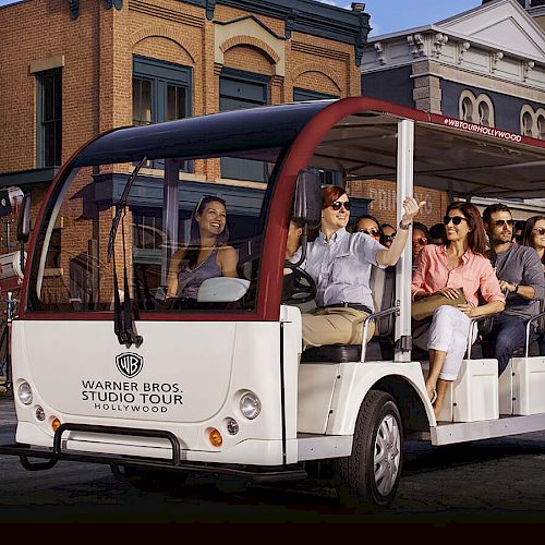 A group of people is seated in an open-air tour vehicle labeled "Warner Bros. Studio Tour Hollywood," passing by a backdrop of building facades.