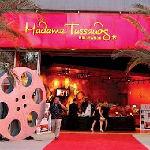 The image shows the entrance to Madame Tussauds Hollywood, featuring a red carpet, palm trees, and large film reel decorations.