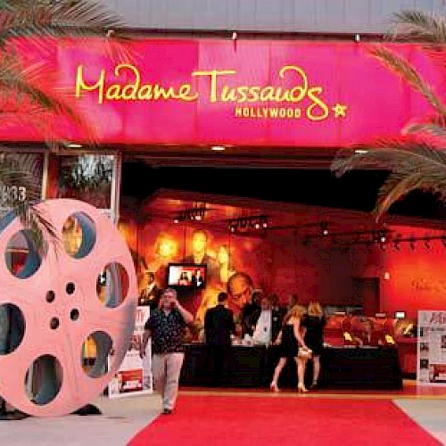 The image shows the entrance to Madame Tussauds Hollywood, featuring a red carpet, palm trees, and large film reel decorations.