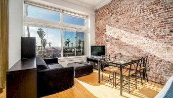 A modern living room with a brick accent wall, large windows, a black sofa, a TV, and a dining table with chairs, featuring a palm view outside.