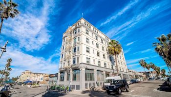 A multi-story building stands on a sunny street corner with palm trees and a bright blue sky. Some vehicles are parked nearby, ending the sentence.