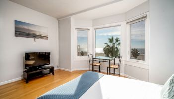 A bedroom with hardwood floors, a bed, a TV, and a small dining table by bay windows with an ocean view, and a wall photo.