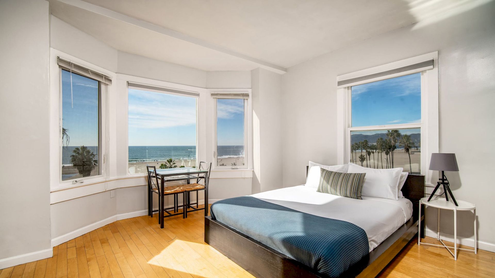 A spacious bedroom with a bed, side table, lamp, small desk, and large windows offering an ocean view with lots of natural light coming in.