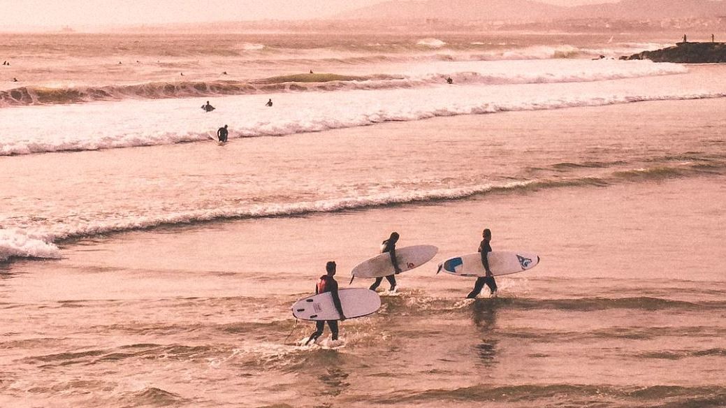 Three people carrying surfboards walk along the shore, with waves and other surfers in the background under a pinkish sky.