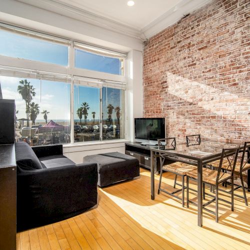A modern loft-style living room with an exposed brick wall, large window view of palm trees, a black sofa, a dining table with chairs, and a small TV setup.