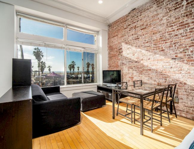A modern loft-style living room with an exposed brick wall, large window view of palm trees, a black sofa, a dining table with chairs, and a small TV setup.