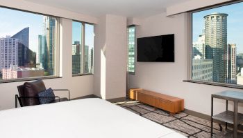 The image displays a modern hotel room with large windows offering views of a city skyline, a mounted TV, and minimalistic furniture.