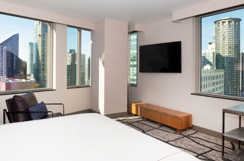The image displays a modern hotel room with large windows offering views of a city skyline, a mounted TV, and minimalistic furniture.
