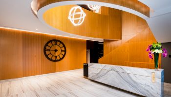 A modern lobby with a marble reception desk, wooden accents, a large wall clock, and decorative ceiling lights.