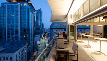 A modern rooftop patio with tables and chairs overlooking a cityscape with high-rise buildings and street below during evening time.