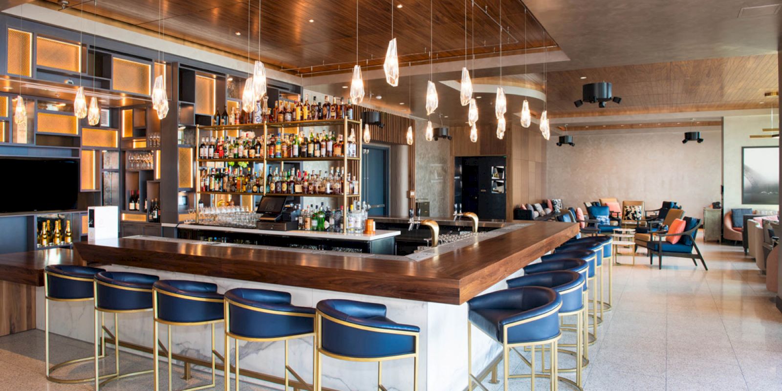 This image shows a modern bar with a sleek wooden counter, blue chairs, hanging pendant lights, and a well-stocked liquor shelf in a stylish interior.