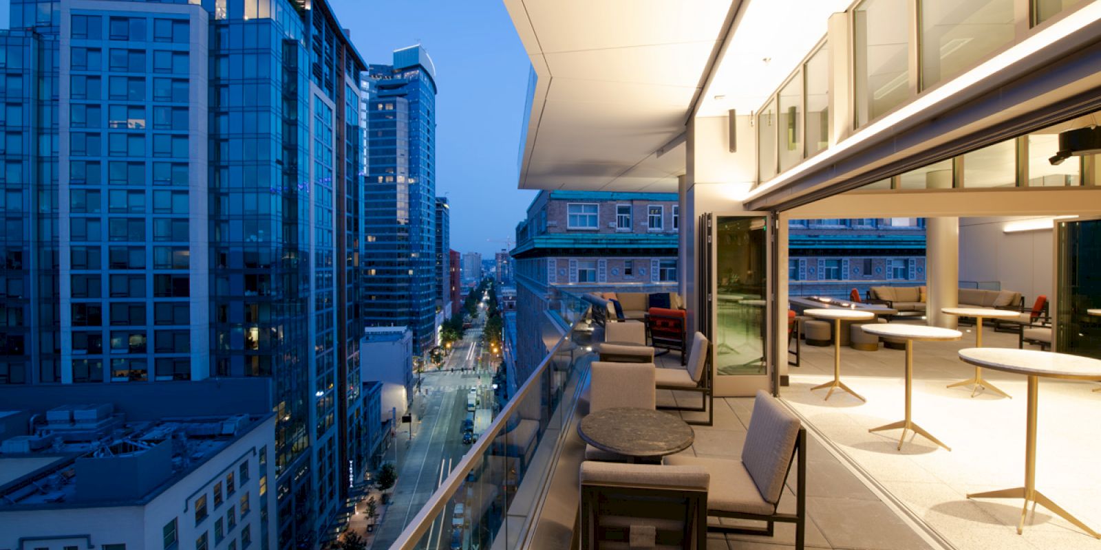A modern cityscape at dusk with tall buildings and a rooftop patio featuring seating and tables on the right-hand side.