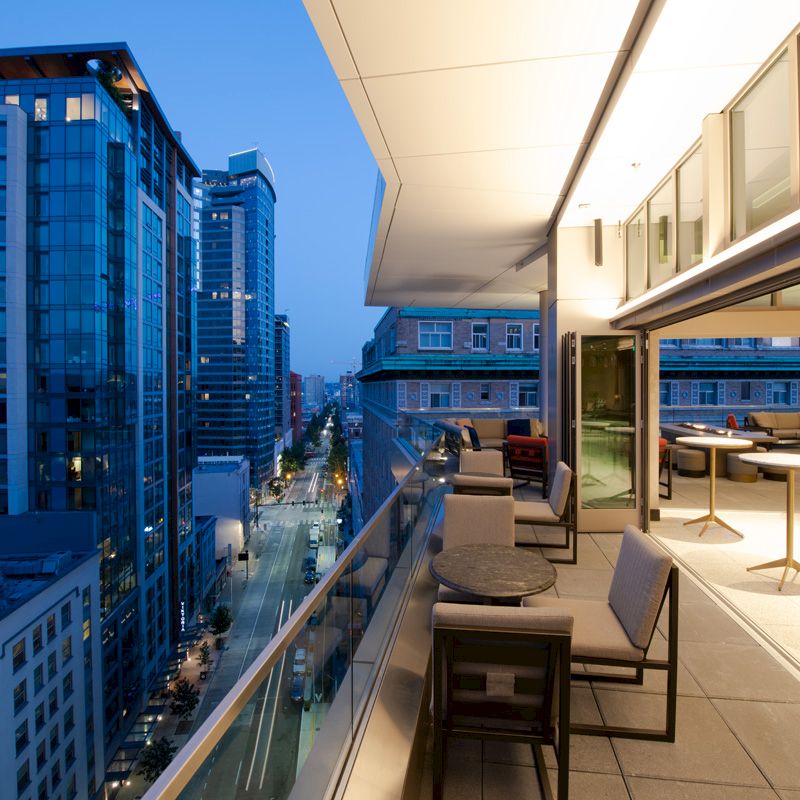 The image shows a modern balcony overlooking a city street with tall buildings at dusk. The balcony has tables, chairs, and bright indoor lights.