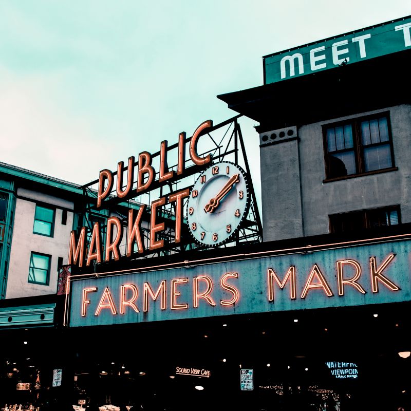 This image shows an iconic Farmers Market sign with a clock, and buildings in the background. The top green sign reads "MEET THE".