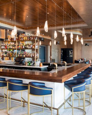 The image shows a modern bar with blue and gold stools, a well-stocked bar, pendant lights, wooden ceiling, and seating areas in the background.