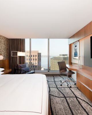 A modern hotel room with a large bed, wall-mounted TV, desk with chair, and a view of buildings and water through the large windows.
