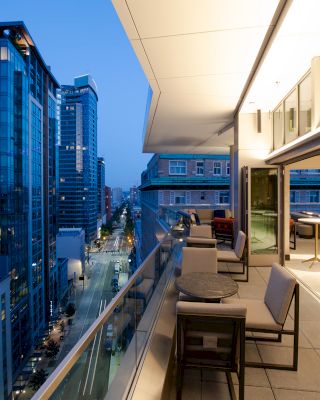 A rooftop terrace in an urban setting at dusk, featuring outdoor seating and a view of modern high-rise buildings along a city street.
