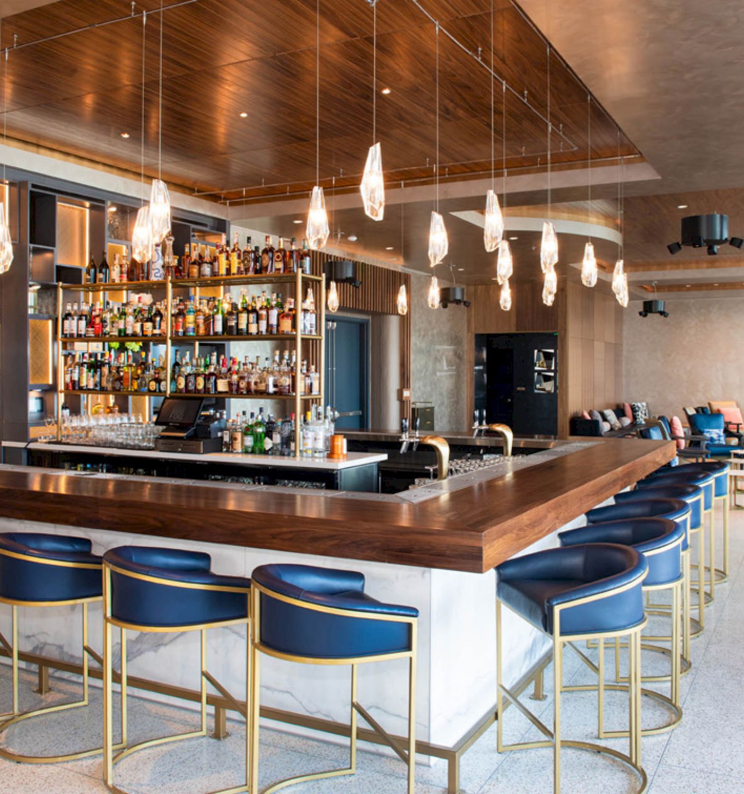 A modern bar with a well-stocked shelf of liquor bottles, blue bar stools, stylish pendant lights, and a cozy seating area in the background.