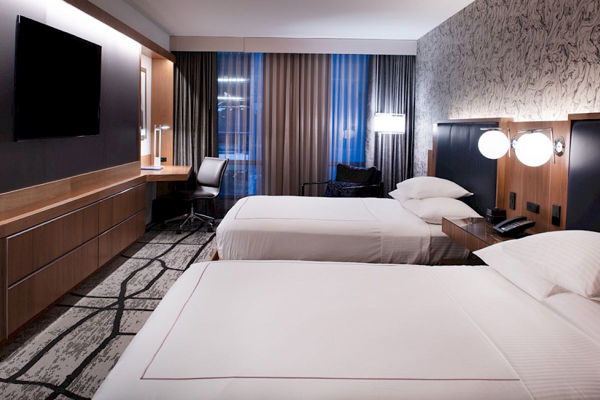 A modern hotel room with two neatly made beds, a wall-mounted TV, a desk with a chair, and contemporary decor.