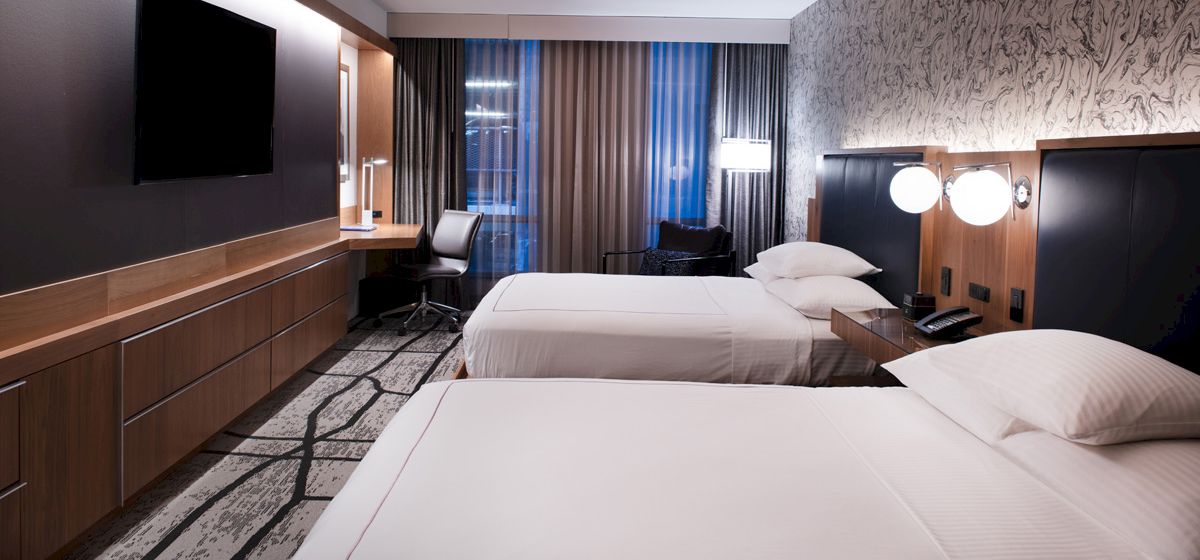 A modern hotel room with two neatly made beds, a wall-mounted TV, a desk with a chair, and contemporary decor.