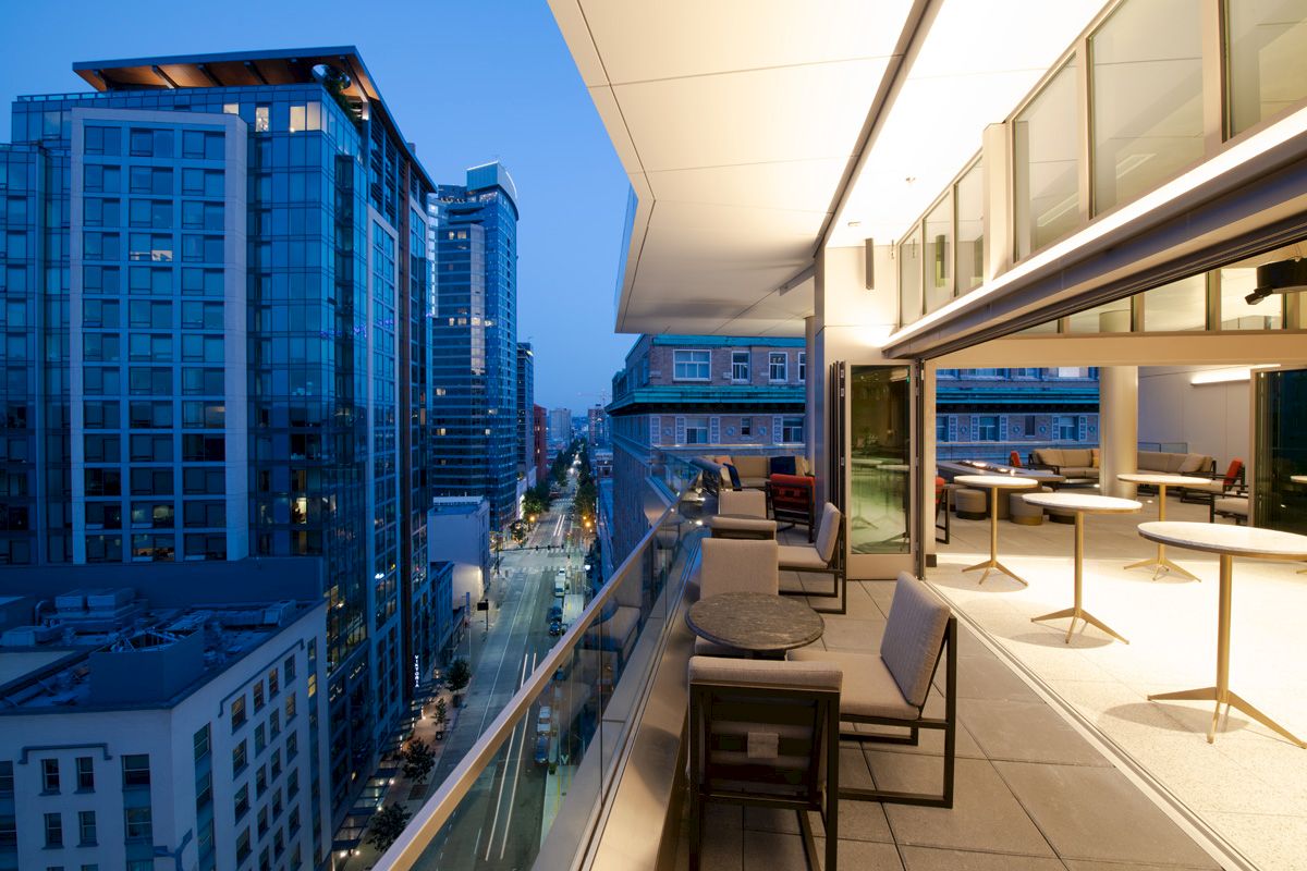 A modern balcony with tables and chairs overlooking a city street lined with high-rise buildings during twilight.