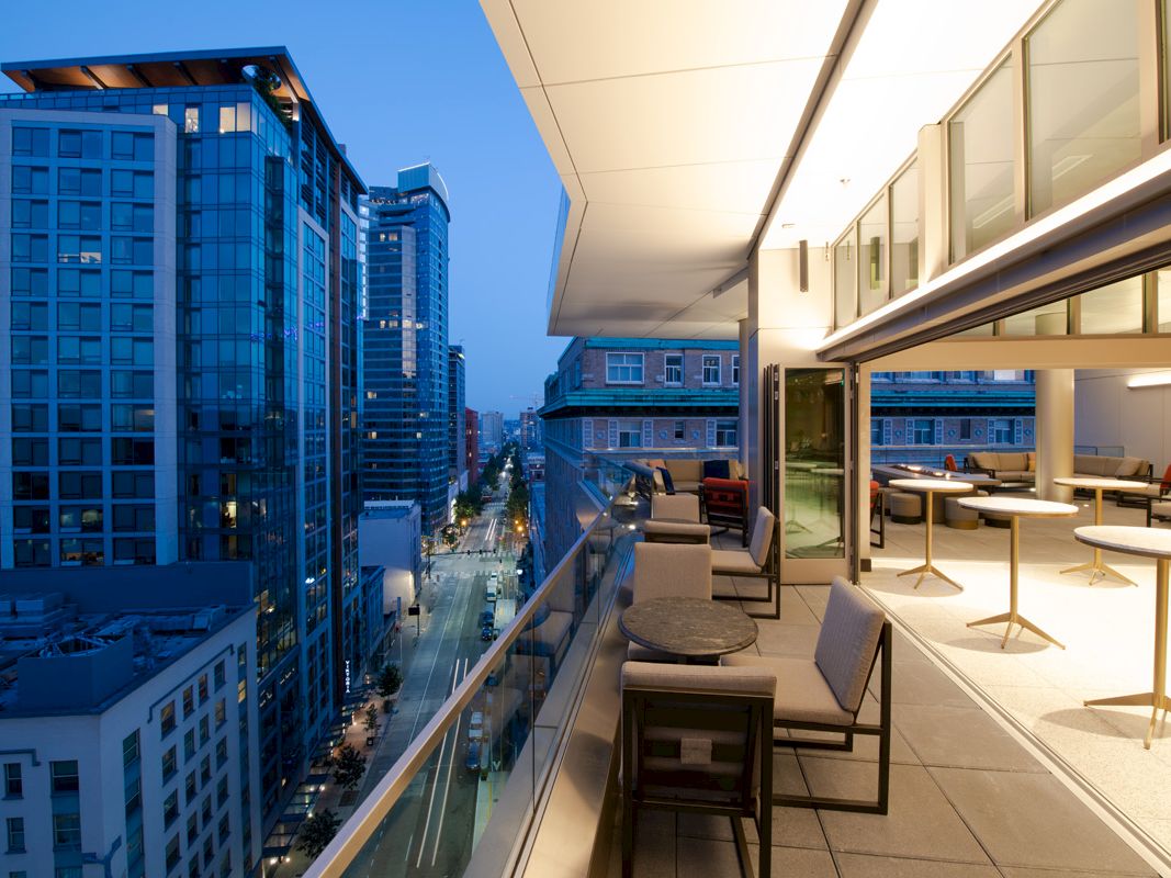 A modern balcony with tables and chairs overlooking a city street lined with high-rise buildings during twilight.