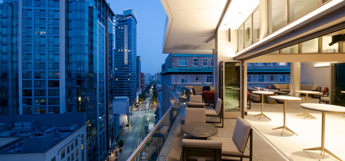 A modern balcony with tables and chairs overlooking a city street lined with high-rise buildings during twilight.