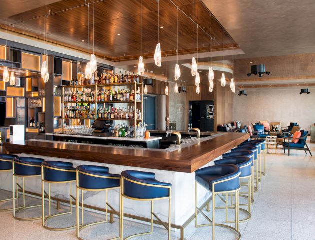 A modern bar with a stocked shelf, stylish pendant lights, and blue bar stools. There are comfortable seating areas in the background.