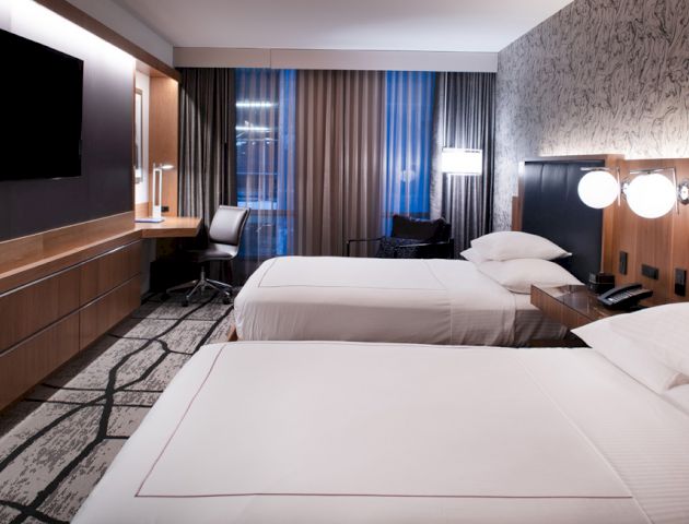 A modern hotel room with two beds, a flat-screen TV, a desk by the window, white linens, and stylish decor on the walls and carpet.