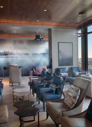 The image shows a modern lounge area with comfortable seating, large windows offering a city and water view, and a wall mural depicting a skyline.