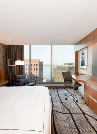 A modern hotel room with a large bed, wall-mounted TV, desk, chair, and large windows offering a view of buildings and the cityscape.