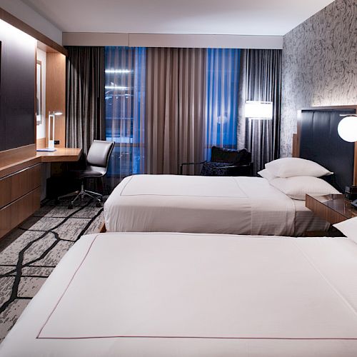 The image shows a modern hotel room with two neatly made beds, a large TV, a desk, and stylish decor, creating a comfortable and inviting atmosphere.