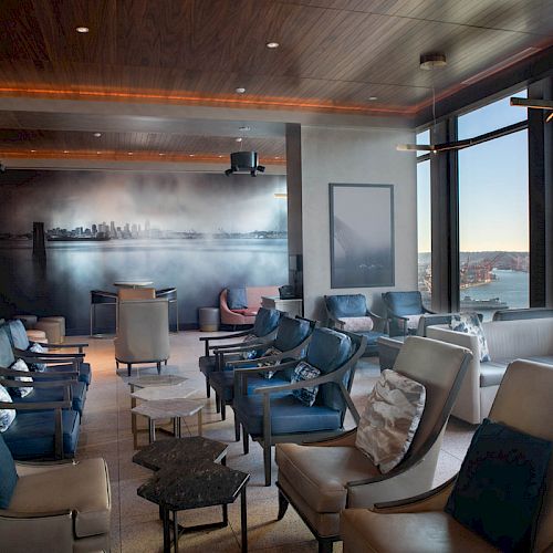 A modern lounge area with comfortable seating, large windows offering a scenic view, and artwork on the wall, overlooking a waterfront scene.