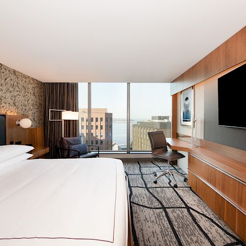 Modern hotel room with a large bed, wall-mounted TV, desk, chair, and a window with an urban and water view in the background.