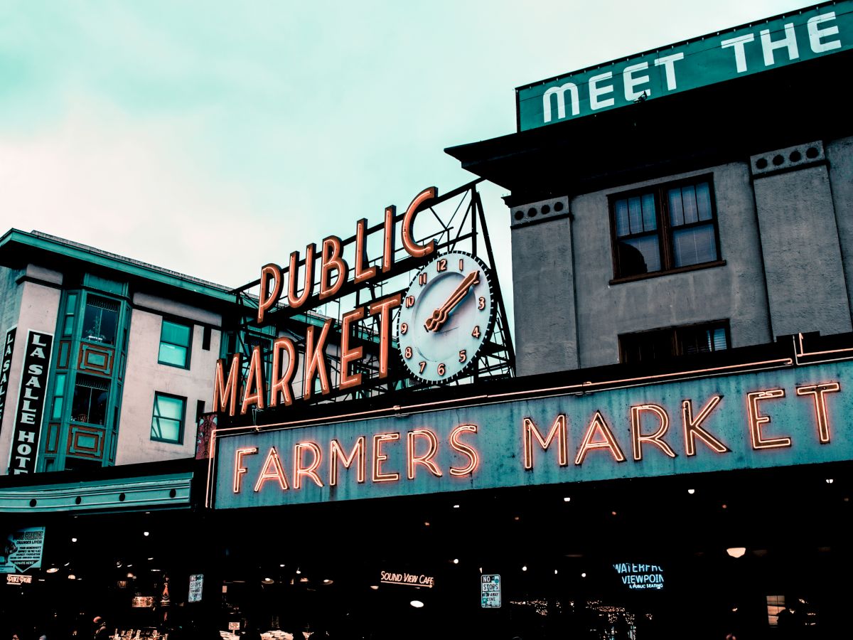 The image shows the exterior of a market building with neon signs that read "PUBLIC MARKET" and "FARMERS MARKET" along with a clock.