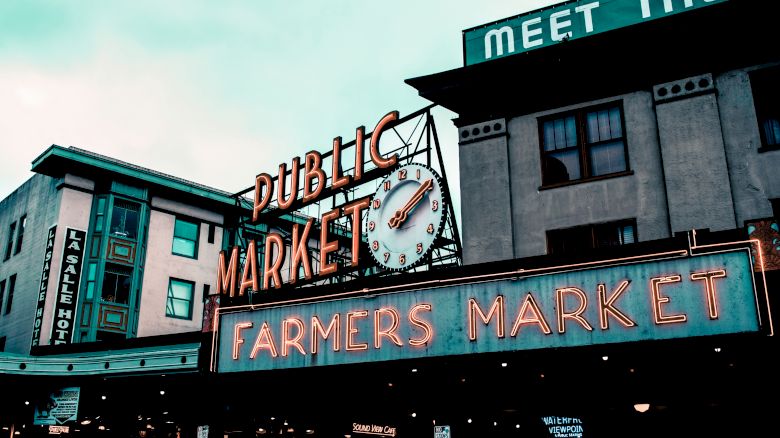 The image shows the exterior of a market building with neon signs that read "PUBLIC MARKET" and "FARMERS MARKET" along with a clock.