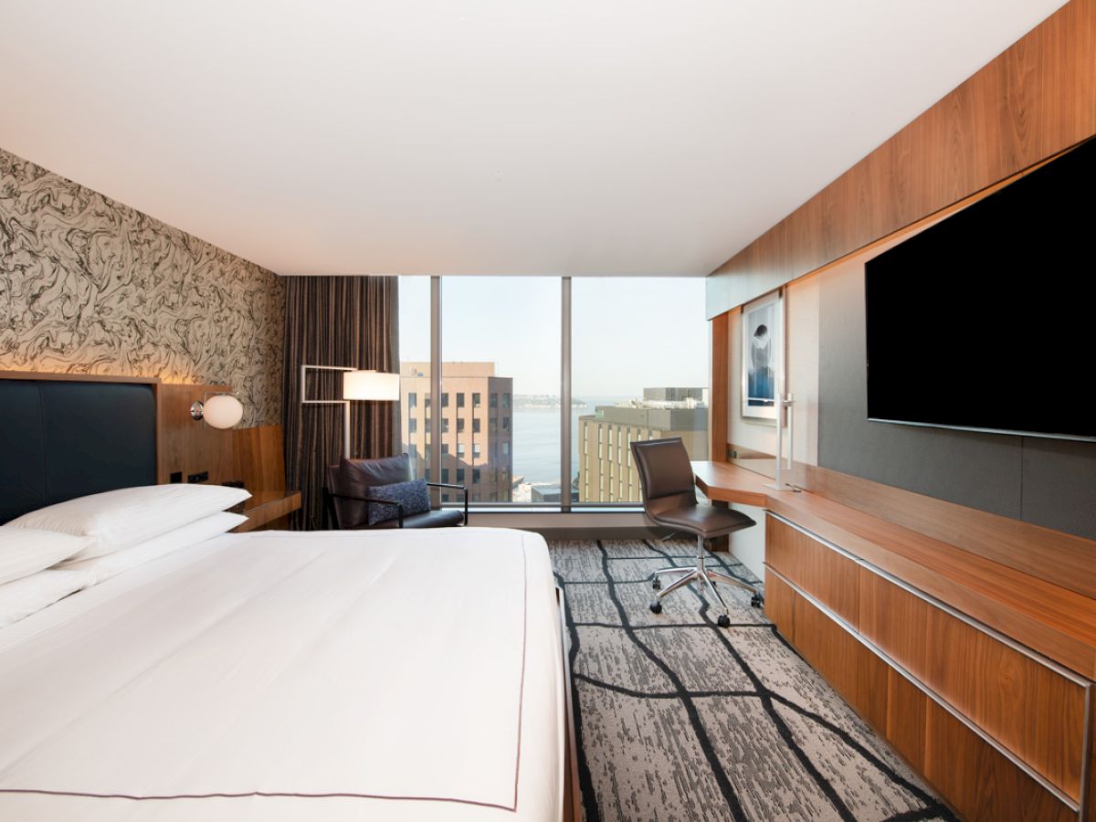 A modern hotel room with a large bed, a desk with a chair, a flat-screen TV, and a wall mural, featuring a window with a cityscape view.