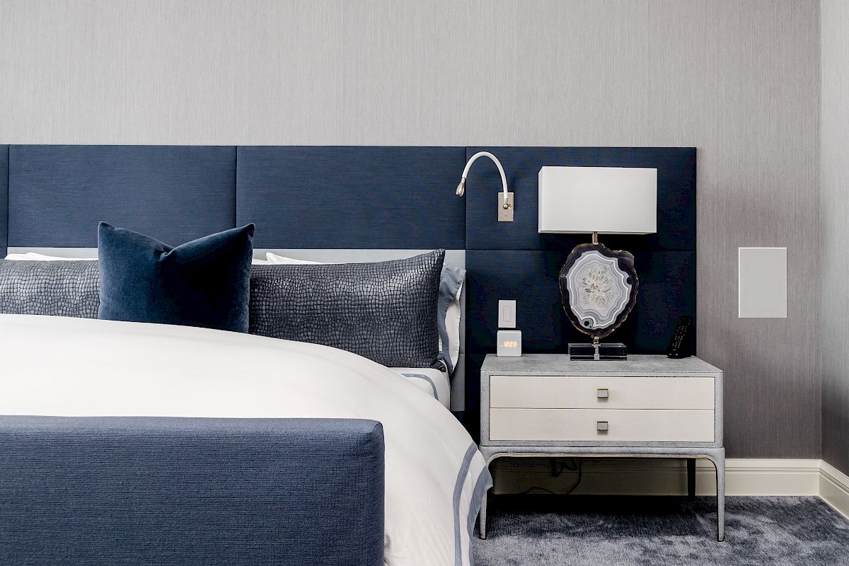 A modern bedroom with a blue upholstered bed, gray and white bedding, a white nightstand, a table lamp, and a wall-mounted reading light.