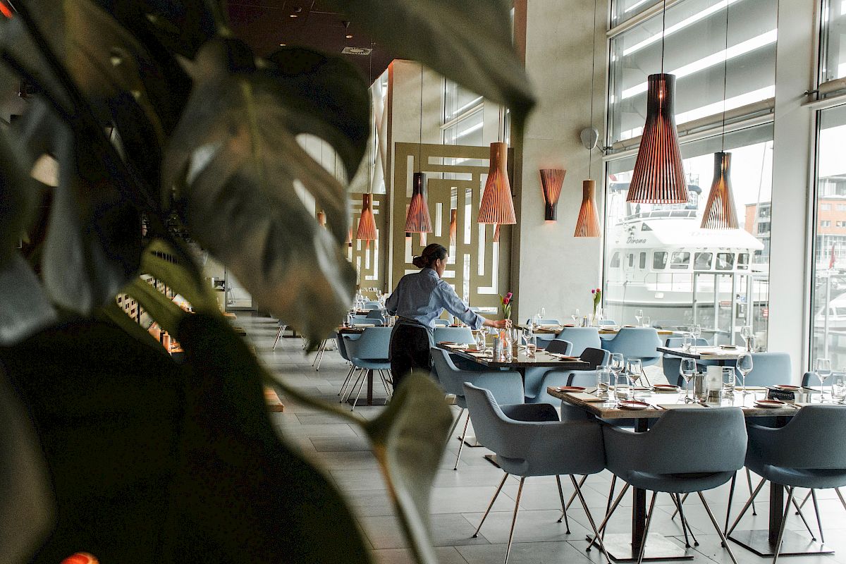 A person is setting tables in a modern, stylish restaurant with large windows and unique lighting fixtures.