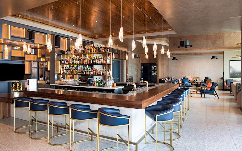 A modern bar with stylish lighting, blue stools, and a wooden countertop is the focal point. Shelves filled with liquor bottles are seen in the background.