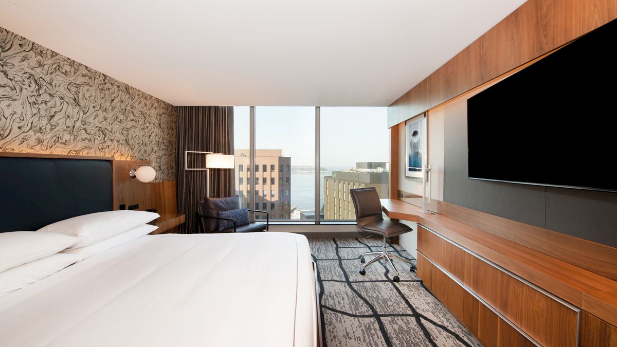 A modern hotel room with a king-size bed, large TV, desk, chair, and a view of buildings outside through a large window.